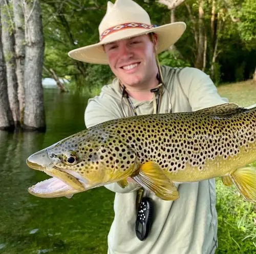 Proud angler holding his catch, a White River Brown Trout, on a beautiful springtime green riverbank.